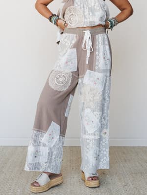 A patchwork wide-leg pant in warm taupe and cream steals the show — layered with crochet doily appliqués, floral embroidery, and lace panels, styled with a matching crop top, stacked bead bracelets, and espadrille platform sandals against a clean stu