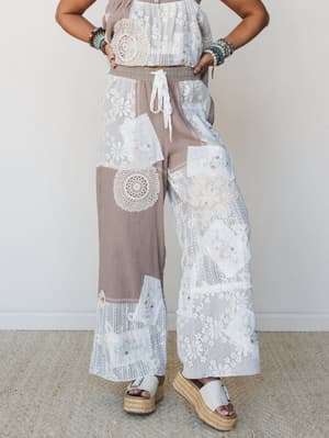 A patchwork wide-leg pant in mocha and white steals the show — mismatched lace panels, crochet doily appliqués, and delicate floral embroidery give it that 'found at a vintage market, but make it fashion' energy, styled with platform espadrille sanda