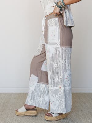 A side-profile shot of wide-leg patchwork pants in warm taupe and cream, layered with lace, crochet, and embroidered floral patches for a vintage-quilt feel — styled with a lace crop top, stacked turquoise bracelets, and espadrille platform sandals.