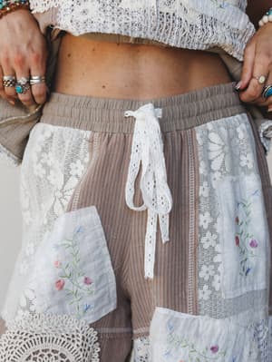 A close-up detail shot of patchwork woven pants layered with lace panels, floral embroidery, and a crochet drawstring waist — styled with stacked silver rings and a cropped lace top, all of it feeling like something stitched together from a grandmoth