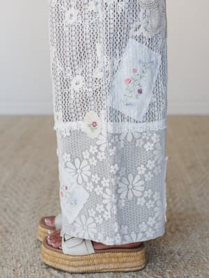 A close-up of wide-leg lace pants with mixed patchwork panels and delicate hand-embroidered floral patches, paired with raffia platform sandals — the kind of piece that looks like it has a whole story behind it.