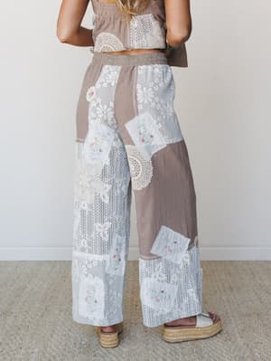 Shot from the back, a model wears wide-leg patchwork pants layered with lace, crochet doilies, and embroidered floral patches over a ribbed chocolate base — paired with a matching cropped top and platform espadrilles, the whole look feels like someth