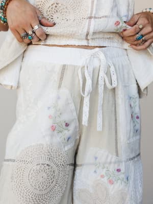 A close-up of cream patchwork wide-leg pants layered with crochet doily patches, delicate floral embroidery, and a lace drawstring waist — paired with a crochet crop top and stacked silver and turquoise rings that feel like they've been collected ove