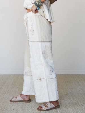 A side-profile shot of a flowy cream patchwork wide-leg pant styled with a matching woven top, turquoise stacked bracelets, and neutral buckle sandals — the kind of outfit that looks like it was assembled from the most beautiful vintage linens you've