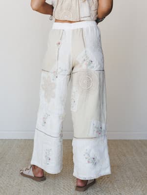 Shot from behind, these wide-leg patchwork pants in cream and natural are a love letter to vintage linens — pieced together with crochet medallions, floral embroidery, and mixed textures that feel like they were made from your grandmother's prettiest