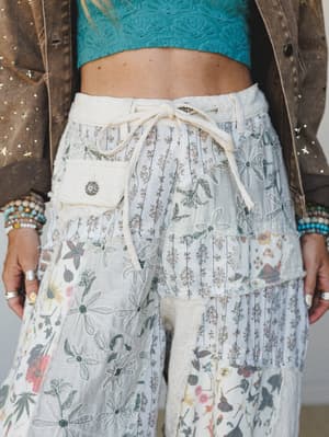 A close-cropped shot of wide-leg patchwork pants in cream and soft blue, layered with mixed floral prints and embroidery, styled with a teal textured crop top and embellished brown jacket — the kind of outfit that looks like it was put together with