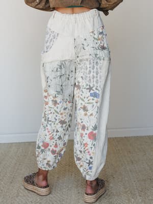 A back-view shot of relaxed, patchwork floral pants in a soft natural cream — mixing botanical prints, embroidered panels, and textured fabrics for that perfectly collected, lived-in feel, styled with platform espadrilles and a cropped olive jacket.