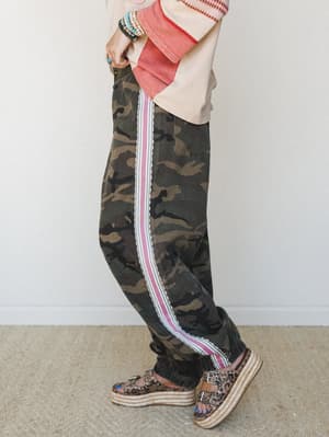 A side-profile shot showing camo wide-leg pants with a bold pink and white side stripe, styled with a washed graphic top, stacked turquoise bracelets, and leopard print platform sandals — the kind of outfit that has a whole personality.