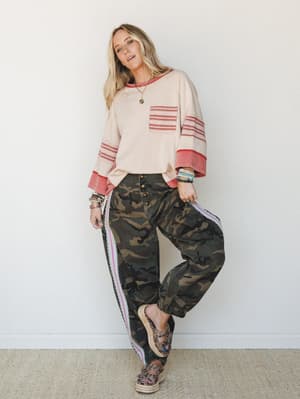 A relaxed full-body shot of a model leaning against a white wall in a cream sweatshirt with red trim and camo jogger pants — the kind of effortlessly thrown-together outfit that somehow looks totally intentional.