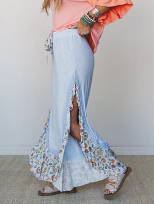 A breezy baby blue maxi skirt-style pant with a flowy floral hem panel and side slit, styled with a coral ruffle tee, stacked boho bracelets, and neutral buckle sandals — the kind of outfit that feels like a slow summer afternoon.