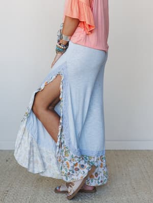 A side-profile shot of a flowy maxi skirt-style wide-leg pant in soft baby blue ribbed fabric, finished with a floral ruffle hem and lace trim, styled with a peach ruffle top and stacked boho bracelets — the kind of outfit that looks effortless but d