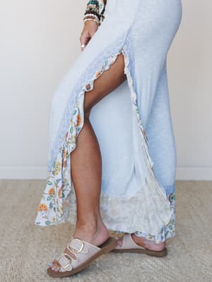 A dreamy close-up of wide-leg pants in soft baby blue with a high slit detail, floral ruffle hem, and lace trim — styled with stacked boho bracelets and buckle slide sandals for that effortlessly romantic, lived-in feel.