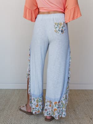 A back-view shot of wide-leg pants in soft baby blue textured fabric, featuring floral ruffle trim at the hem and sides, a floral contrast pocket, and lace trim detailing — paired with a coral knotted tee for an easy, expressive summer look.
