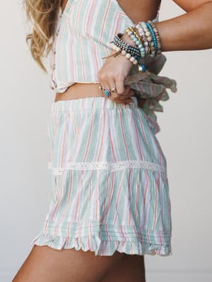 A close-up torso shot of a matching striped two-piece set — a cropped top and ruffle-hem shorts — styled with a stacked arm of beaded bracelets and a turquoise ring, giving off easy summer energy with a soft boho finish.