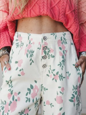 A close-up shot of high-waisted floral pants in cream with pink blooms and sage green leaves, styled with a coral cable-knit cropped sweater and stacked bracelets — the kind of outfit that looks like it was pulled together effortlessly but definitely