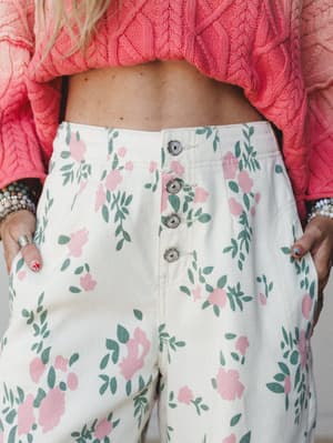 A close-up shot of high-waisted floral pants in cream with pink blooms and sage green leaves, styled with a coral cable-knit cropped sweater and stacked bracelets — the kind of outfit that looks like it was pulled together effortlessly but definitely