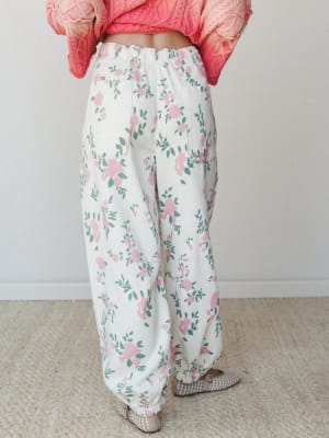 A back-view shot of wide-leg floral pants in cream with pink roses and sage green leaves, paired with a cropped coral knit sweater — the kind of outfit that makes a Tuesday feel intentional.