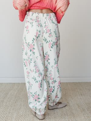 A back-view shot of wide-leg floral pants in cream with pink roses and sage green leaves, paired with a cropped coral knit sweater — the kind of outfit that makes a Tuesday feel intentional.