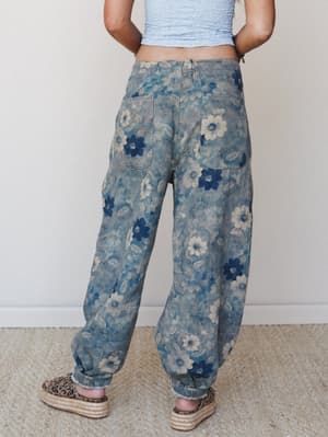 A back-view shot of relaxed, wide-leg jogger pants in a washed floral print — navy and cream daisy blooms over a faded blue-grey base — styled with a cropped smocked top and platform espadrilles for an effortlessly boho everyday look.