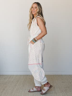 A full-length cream jumpsuit with a ditsy floral print, peace sign appliqué on the leg, and red contrast stitching — worn with stacked boho bracelets and Birkenstock-style sandals, styled against a clean neutral wall for an easygoing, free-spirited f