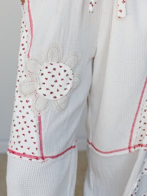 A close-up detail shot of a cream mixed-media jumpsuit with charming patchwork panels — ditsy floral print fabric, waffle knit texture, red contrast stitching, and a crochet flower appliqué that feels like something your grandmother's quilt and your