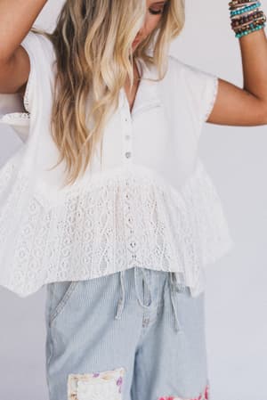 A woman with wavy blonde hair wears the Callie Short Sleeve Tee in white and light blue striped patchwork shorts, accessorized with colorful beaded bracelets. Her arms are raised and her face is partially out of frame.