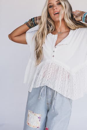 A woman with long blonde hair wears the Callie Short Sleeve Tee in white, paired with light blue striped patchwork pants and colorful beaded bracelets, standing against a plain light background.
