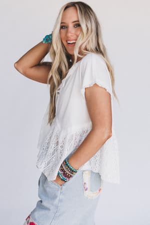 A woman with long blonde hair smiles, standing sideways. She wears the Callie Short Sleeve Tee in white, light blue striped pants with colorful patches, and multiple beaded bracelets. The background is plain white.