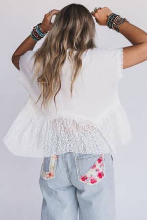 A woman with long, wavy blonde hair faces away, wearing the Callie Short Sleeve Tee in white and light blue jeans with floral patches. She lifts her arms, showing off colorful beaded bracelets on both wrists.