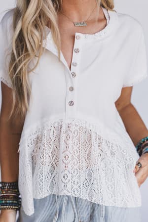 A woman wears the Callie Short Sleeve Tee in white, featuring a lace trim hem and buttoned neckline. She styles it with layered necklaces and colorful bracelets, her long blonde hair down. Only her upper body is visible.