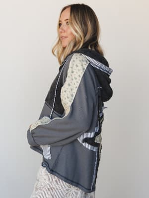 A model shot in three-quarter profile wearing a patchwork hoodie in charcoal waffle knit with mixed fabric panels — floral, stripe, and sherpa — layered over a striped skirt, giving it that collected, one-of-a-kind feeling.