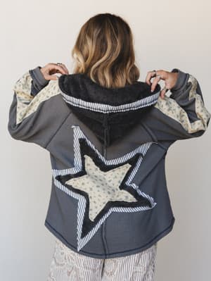 A model shown from behind pulls up the sherpa-lined hood of a charcoal waffle-knit jacket, revealing a layered star appliqué on the back built from striped fabric, floral print, and raw-edge fleece detailing — the kind of piece that makes someone sto