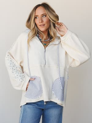 A relaxed, mixed-media pullover hoodie in cream waffle knit with blue stripe and ditsy floral patchwork panels, styled with jeans and layered beaded necklaces — the kind of top that looks intentional without trying too hard.