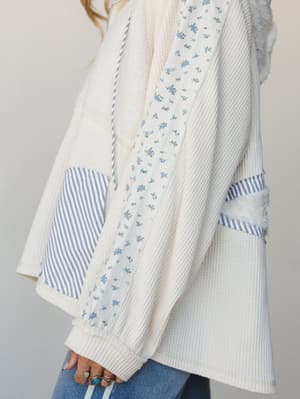 A close-up side view of a cream waffle-knit pullover with charming patchwork detailing — ditsy floral fabric panels, blue and white stripe pocket and sleeve accents, and a drawstring hood, styled with denim and stacked silver rings.