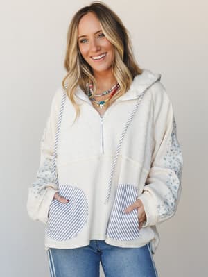 A cream waffle-knit zip-up hoodie with mixed fabric detailing — floral print sleeves and striped kangaroo pockets give it that intentionally patchwork, lived-in feel, styled with denim and layered bead necklaces for an easy, expressive everyday look.