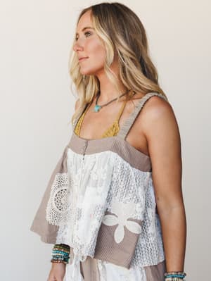 A side-profile shot of a model wearing a relaxed, patchwork tank top in warm taupe ribbed cotton with mixed white lace and crochet panels — layered over a mustard lace bralette and styled with stacked turquoise and beaded bracelets and a turquoise pe