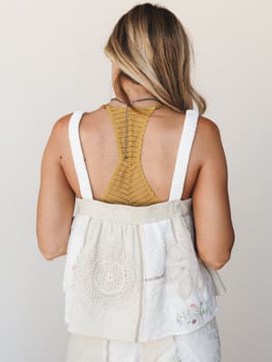 A back-facing shot of a model wearing a cream woven patchwork top with crochet medallion and floral embroidery details, layered over a mustard lace racerback bralette — effortlessly boho with just the right amount of texture.
