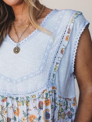 A close-up detail shot of a baby blue mixed-fabric top with delicate crochet trim and a floral ruffle hem, styled with layered gold necklaces — the kind of piece that feels like a farmers market morning and a backyard dinner all in one.