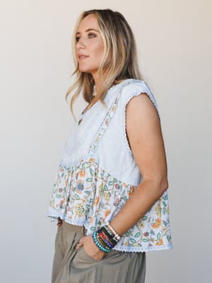 A breezy mixed-fabric top in baby blue pairs a textured solid bodice with a floral printed hem panel, finished with crochet lace trim — styled with olive trousers and stacked beaded bracelets for an effortlessly personal, warm-weather feel.