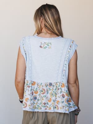 A back view of a mixed-media peplum top in baby blue — textured gauze bodice meets a flowy floral hem panel, held together with delicate lace trim throughout.