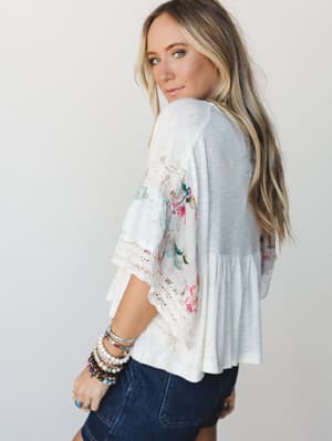 A model glances back over her shoulder in a cream mixed-media peplum top — waffle-knit back panel meets floral-print sleeves with lace trim detailing, paired with dark denim and a stack of boho bracelets that feel entirely personal.