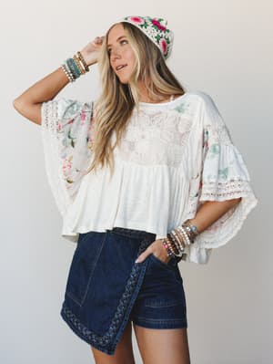A breezy, layered boho look built around a flowy white patchwork top with floral sleeves and lace panels, paired with dark denim braided-trim shorts and a crochet floral beanie — the kind of outfit that looks effortless but definitely took some thoug