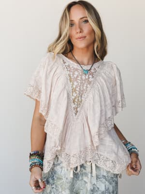 A flowy blush lace-trimmed top with an embroidered sheer V-neckline panel sits softly on the model, styled with stacked boho bracelets and a turquoise pendant — the kind of top that looks like it was made for slow mornings and good light.