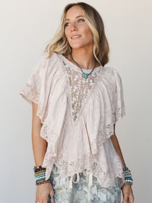A flowy, layered blouse in soft blush almond with a sheer floral embroidered center panel and crochet lace trim throughout — styled with tie-dye shorts and stacked boho bracelets for an effortlessly romantic look.