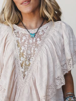 A dreamy close-up of a blush linen-look blouse with a sheer floral embroidered yoke and crochet lace trim throughout — styled with a turquoise butterfly pendant and stacked bracelets for that effortless boho-romantic vibe.