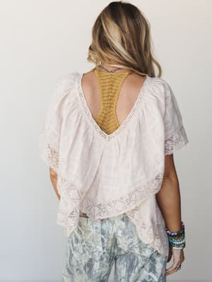 Shot from behind, this flowy lace-trimmed blouse in soft almond shows off a deep v-back with a mustard yellow bralette peeking through — styled with floral-print pants and a stack of beaded bracelets for a fully lived-in boho moment.