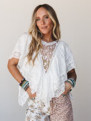 A breezy white lace-trimmed blouse with a sheer floral embroidered panel styled with mixed-print bottoms and stacked boho bracelets — the kind of top that makes a casual Tuesday feel intentional.