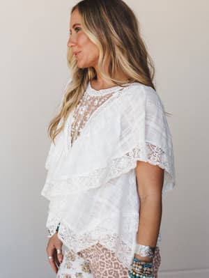A flowy white lace blouse with tiered ruffle layers and delicate embroidered lace trim is styled with stacked boho bracelets and a leopard-print skirt peeking through — effortless and a little romantic.
