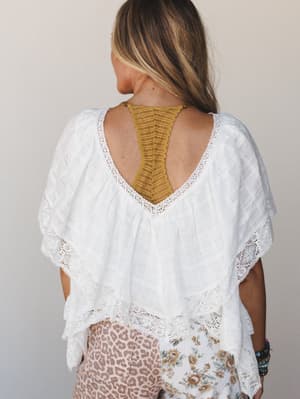 Shot from behind, this breezy white lace-trim blouse has a deep V-back that shows off a mustard yellow bralette underneath — effortlessly boho, the kind of top that does all the work while feeling like nothing.