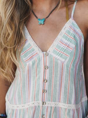 A close-up detail shot of a striped button-front cami top with lace trim, styled with a turquoise butterfly pendant necklace — soft, boho, and exactly the kind of thing you'd reach for on a warm afternoon.